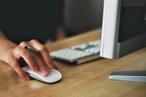 Hand holding an Apple Magic Mouse beside a keyboard, symbolizing precise digital workflows and clean, validated product data managed through a PIM system.